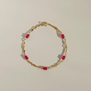May include: A gold-coloured bracelet with a series of red beads interspersed with elongated gold links. The bracelet, with its simple clasp, is presented against a plain background. A stylish piece of jewellery.