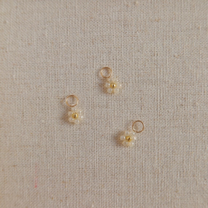 Op de afbeelding: Drie goudkleurige bedels met witte parel-madeliefjes. De bedels hebben een kleine lus aan de bovenkant om aan sieraden te bevestigen.