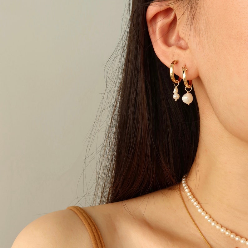 May include: Gold hoop earrings with a pearl drop dangle. The earrings are worn by a person with long dark hair.