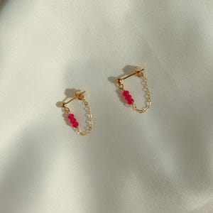 Può includere: Un paio di orecchini color oro con una catena e tre piccole perline rosse. Gli orecchini sono presentati su una superficie bianca, leggermente strutturata. Design con perno e catena.