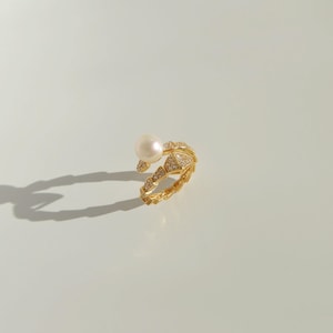 May include: A gold-colored ring with a snake-like design, featuring a large white pearl and small, sparkling stones. The ring is set against a white background, casting a shadow.