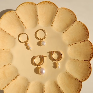 May include: Three pairs of gold hoop earrings with pearl drops. The earrings are displayed on a yellow scalloped plate.
