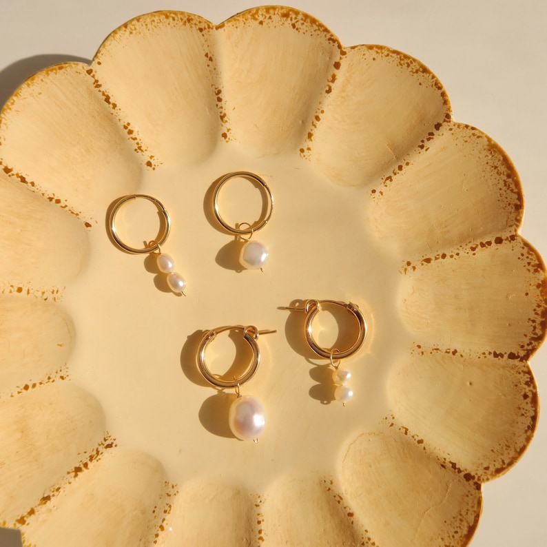 May include: Three pairs of gold hoop earrings with pearl drops. The earrings are displayed on a yellow scalloped plate.