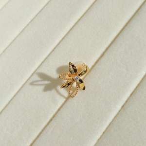 May include: A gold-colored floral stud earring with a small, clear gemstone at the center. The earring has a delicate, openwork design with a pearl-like accent. The earring is set against a cream-colored, textured background.