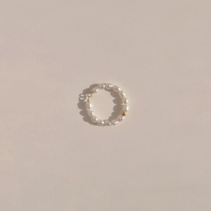 May include: A delicate ring featuring a circle of white pearls, accented with a small gold-coloured bead and a gold-coloured clasp. The ring is set against a neutral background, highlighting its elegant design. This piece of jewellery is perfect for adding a touch of sophistication.