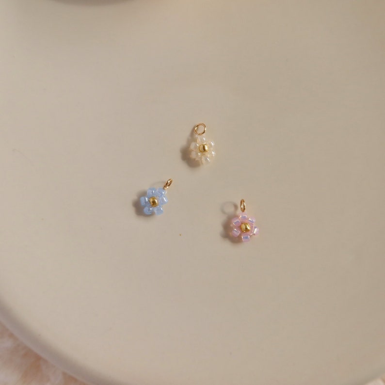 Op de afbeelding: Drie kleine bloemcharms gemaakt van kralen. De charms zijn lichtblauw, wit en roze. Elke charm heeft een gouden middelste kraal.