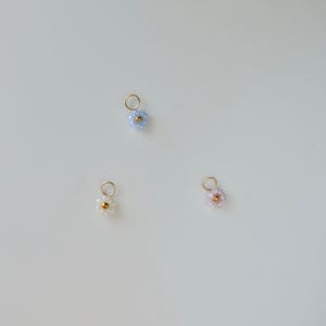 Op de afbeelding: Drie kleine kralenbloembedels in blauw, wit en roze. Elke bedel heeft een gouden middelste kraal en is bevestigd aan een gouden springring.