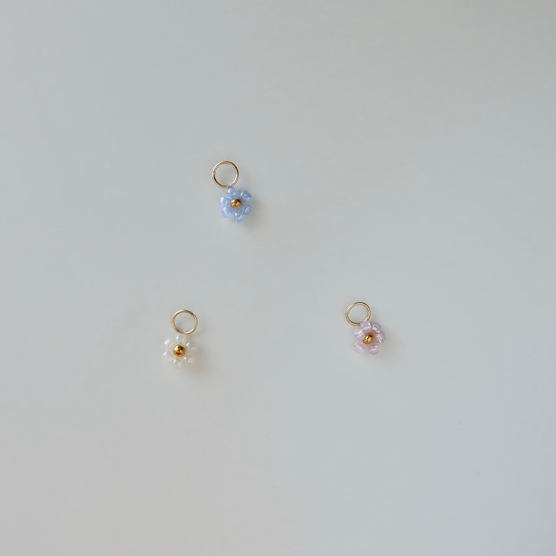 Op de afbeelding: Drie kleine kralenbloembedels in blauw, wit en roze. Elke bedel heeft een gouden middelste kraal en is bevestigd aan een gouden springring.