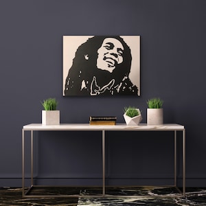 Può includere: Dipinto in silhouette in bianco e nero di Bob Marley su una tela bianca, appeso a una parete grigio scuro sopra un tavolo consolle in marmo bianco con due piante in vaso e una pila di libri.