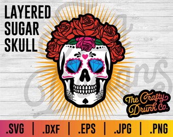 May include: A white sugar skull with blue and pink details, wearing a wreath of red roses. The skull has a heart on its forehead and diamond-shaped eyes. The text "The Crafty Drunk Co." is in the lower right corner of the image.