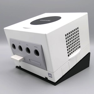 Może przedstawiać: Biała konsola Nintendo GameCube na czarnej podstawie. Konsola ma okrągły przycisk zasilania i otwór wentylacyjny z boku. Z przodu znajdują się porty kontrolera i gniazdo karty pamięci.
