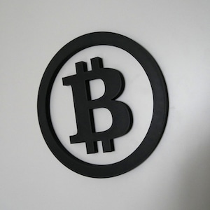 Puede incluir: Un símbolo de Bitcoin en blanco y negro en un círculo negro sobre una pared blanca.