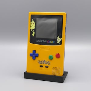 Puede incluir: Una consola de videojuegos portátil Game Boy Color amarilla con una placa frontal negra y azul. La consola tiene una pantalla, botones y un altavoz. La consola está en un soporte negro. La consola tiene un logotipo de Pokemon y el texto "GAME BOY COLOR" en la parte delantera.