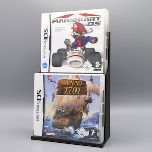 Könnte beinhalten: Zwei Nintendo DS-Spieleboxen auf einem schwarzen Ständer. Die obere Box ist für Mario Kart DS und zeigt einen weißen Hintergrund mit Mario, der ein Kart fährt. Die untere Box ist für Anno 1701 und zeigt ein Schiff, das auf dem Meer segelt.