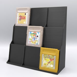 Könnte beinhalten: Schwarzer Game Boy-Cartridge-Displayständer aus Kunststoff mit drei Fächern. Der Ständer hält drei Game Boy-Cartridges, eine mit einer goldenen Cartridge, eine mit einer beigefarbenen Cartridge und eine mit einer beigefarbenen Cartridge. Die Cartridges haben die Titel "Pokemon", "Super Mario Land 2" und "The Legend of Zelda: Link's Awakening".
