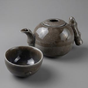 May include: A glossy, dark grey ceramic teapot with a rounded body, a curved spout, and a textured handle shaped like a branch, accompanied by a matching small, round, dark grey ceramic tea cup with a smooth, reflective interior surface.