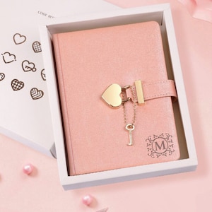 May include: A pink leather-bound journal with a heart-shaped lock and key. The journal has a gold-colored initial "M" on the front cover. The journal is in a white box with a pink interior.