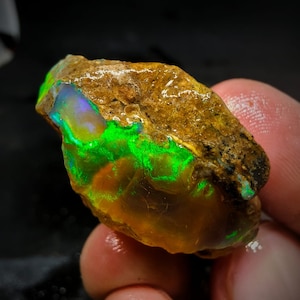 Pode incluir: Um opala natural bruto com um brilho verde e iridescente. O opala está engastado em uma matriz marrom.