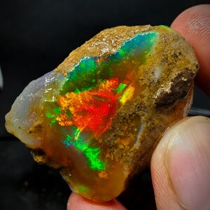 Pode incluir: Uma rocha áspera, marrom e cinza com um grande opala iridescente dentro. O opala exibe um arco-íris de cores, incluindo vermelho, laranja, amarelo, verde, azul e roxo.