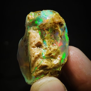 Pode incluir: Um opala natural bruto com um brilho iridescente verde, azul e vermelho. O opala está engastado em uma matriz de rocha marrom e branca.