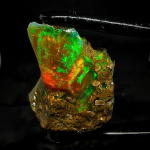 Pode incluir: Uma gema de opala bruta, verde e laranja, com uma matriz marrom. A opala é iridescente e tem um brilho verde brilhante.