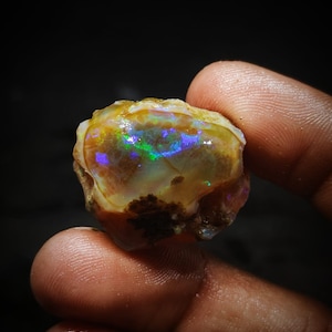 Pode incluir: Uma gema de opala natural bruta com um exterior marrom, branco e verde. O interior da opala exibe uma vibrante exibição iridescente de verde, azul e roxo.