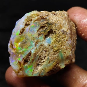 Pode incluir: Um opala natural bruto com uma matriz marrom. O opala exibe um arco-íris de cores, incluindo verde, azul e amarelo.