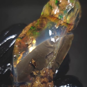 Pode incluir: Um close-up de uma pedra preciosa de opala bruta. A opala exibe uma mistura de cores, incluindo verde, laranja e azul, com uma qualidade translúcida. A pedra bruta tem uma superfície texturizada e está em um fundo escuro.