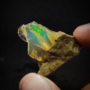 Pode incluir: Uma rocha marrom áspera com um grande cristal de opala iridescente. A opala exibe um arco-íris de cores, incluindo verde, azul e amarelo.