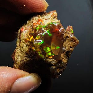 Pode incluir: Uma rocha áspera, marrom e cinza com um pedaço de opala iridescente. A opala exibe flashes de verde, vermelho e laranja.