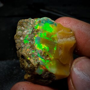 Pode incluir: Um opala natural bruto com um brilho iridescente verde e laranja. O opala está incrustado em uma matriz rochosa marrom.