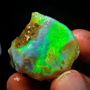 Pode incluir: Uma gema de opala bruta, verde e azul, com uma base branca. A opala é iridescente e tem uma textura áspera e natural.