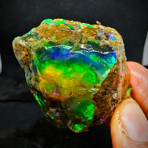 Pode incluir: Um opala natural bruto com um brilho iridescente verde, azul e amarelo. O opala está engastado em uma matriz de rocha marrom.
