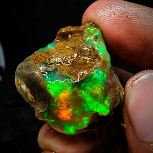 Pode incluir: Um opala natural bruto com um brilho iridescente verde e laranja. O opala está engastado em uma matriz marrom.