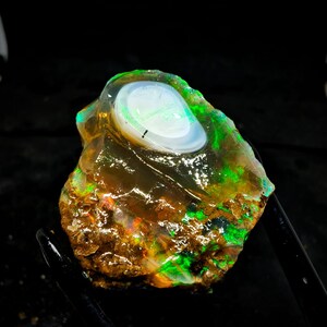 Pode incluir: Um opala natural bruto com um brilho iridescente verde e branco. O opala está engastado em uma matriz marrom.