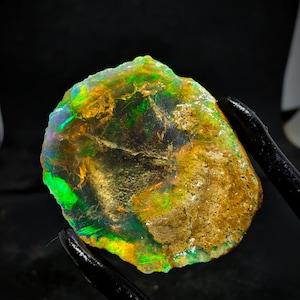 Pode incluir: Uma gema de opala natural bruta com um brilho iridescente verde, amarelo e laranja. A opala tem uma forma irregular e uma matriz marrom terrosa.