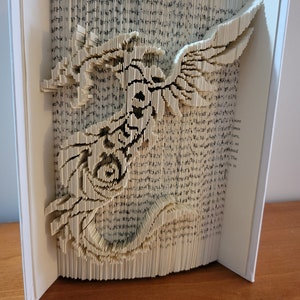 May include: A book art sculpture of a dragon made by folding the pages of a book. The dragon is in flight with its wings spread wide. The sculpture is made from a book with white pages and black text.