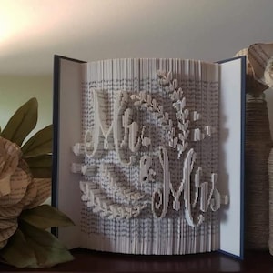 May include: A handcrafted book art piece with the words "Mr & Mrs" cut into the pages. The book is open, revealing the sculpted text and floral accents. The edges of the pages are visible, and the book is set against a neutral background.