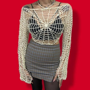 Pode incluir: Um top branco de crochê com um design de teia de aranha. O top é curto e tem mangas compridas. A modelo está usando um sutiã preto por baixo do top.