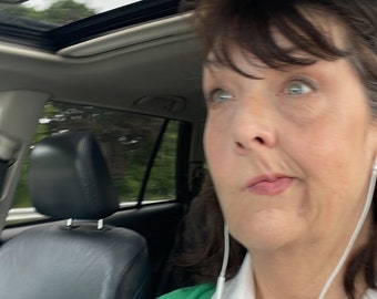 Può includere: Una donna indossa un cardigan verde sopra una camicia bianca con colletto e una collana di perle verdi. Indossa auricolari bianchi e ha una cintura di sicurezza nera allacciata sul petto.