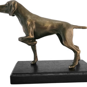 Op de afbeelding: Een bronzen beeld van een Duitse Korthaar, staand op een zwarte houten voet. De hond staat in een jachthouding met zijn voorpoot omhoog.