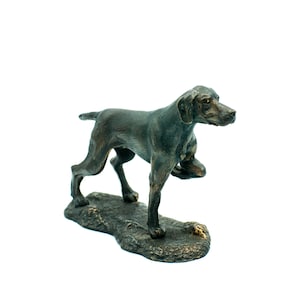 Könnte beinhalten: Eine Bronzeskulptur eines Hundes, möglicherweise eines Pointers, der auf einem felsigen Sockel steht. Der Hund ist in einer Jagdstellung, mit dem Kopf zur Seite gedreht und der rechten Pfote erhoben.