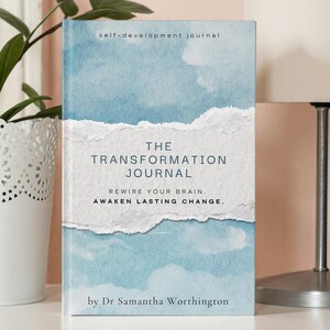 May include: A self-development journal titled "The Transformation Journal" with a blue and white watercolour design. The book is next to a white lamp and a white decorative plant pot. The text on the book reads "Rewire Your Brain, Awaken Lasting Change."