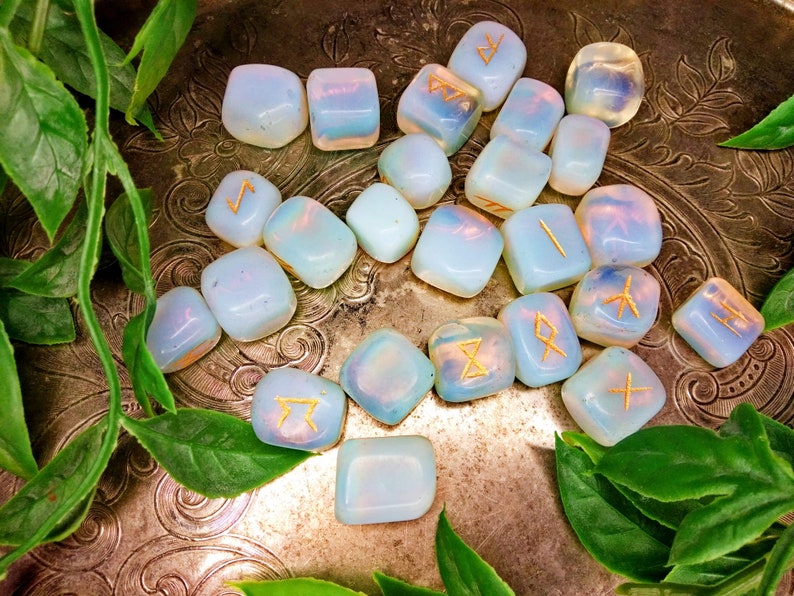 May include: A collection of opalescent, square-cut rune stones with gold-colored symbols. The stones are arranged on a decorative, silver-toned metal surface, surrounded by green leaves. The runes are likely used for divination or spiritual practices.