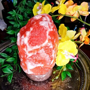 以下が含まれることがあります： ピンクと白の色合いの、大きく、おおまかに形作られたヒマラヤ岩塩ランプ。まだらのベースの上に置かれています。 ランプは黄色の蘭と緑の葉に囲まれ、装飾的な銀色のトレイに置かれています。