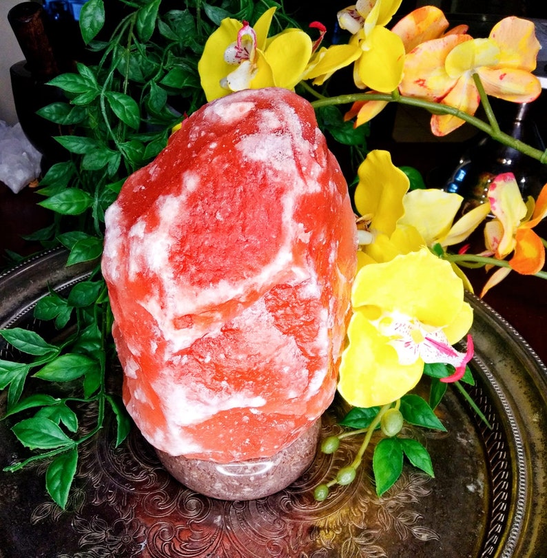 以下が含まれることがあります： ピンクと白の色合いの、大きく、おおまかに形作られたヒマラヤ岩塩ランプ。まだらのベースの上に置かれています。 ランプは黄色の蘭と緑の葉に囲まれ、装飾的な銀色のトレイに置かれています。
