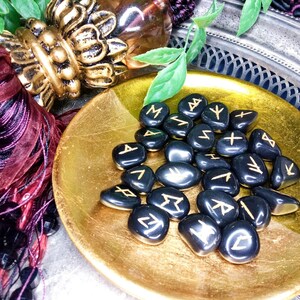 May include: A collection of black rune stones with gold-colored symbols, displayed in a gold-colored, shallow bowl. The bowl sits on a silver tray, with decorative burgundy ribbons and beads in the background. The stones are smooth and polished.