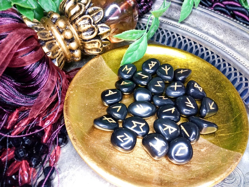 May include: A collection of black rune stones with gold-colored symbols, displayed in a gold-colored, shallow bowl. The bowl sits on a silver tray, with decorative burgundy ribbons and beads in the background. The stones are smooth and polished.