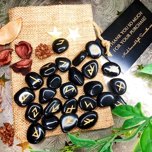 May include: A collection of black, polished rune stones with gold-colored symbols, displayed on a burlap bag. The image includes a small tag with the text "THANK YOU SO MUCH FOR YOUR PURCHASE!" and decorative elements.