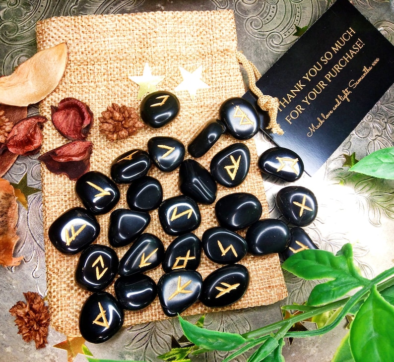 May include: A collection of black, polished rune stones with gold-colored symbols, displayed on a burlap bag. The image includes a small tag with the text "THANK YOU SO MUCH FOR YOUR PURCHASE!" and decorative elements.